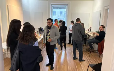People stand and talk in small groups in a bright room with a wooden floor and a tall window.